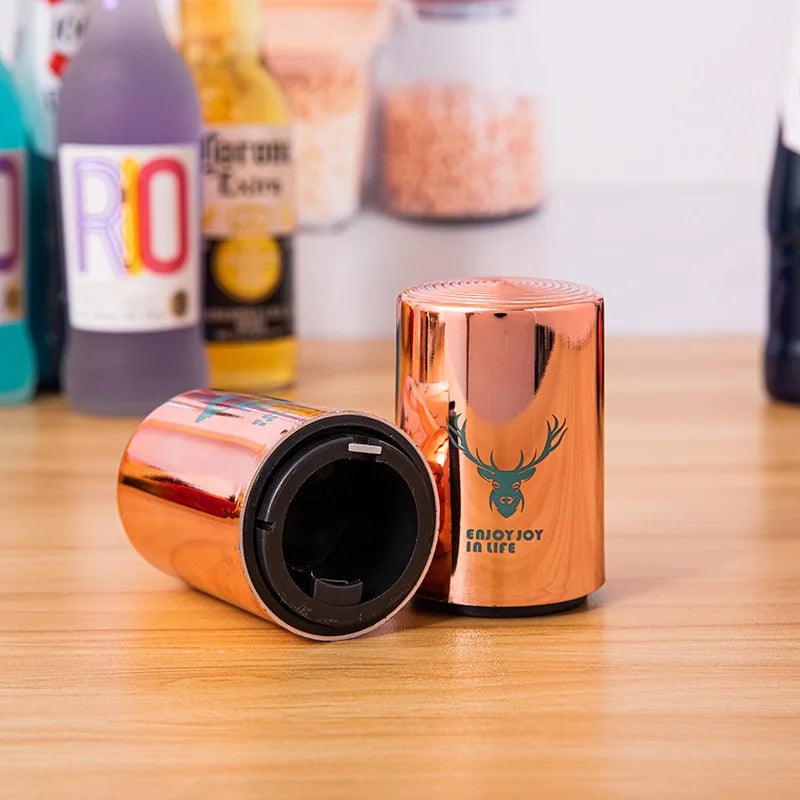 Two copper can openers with robot and deer designs on a wooden surface, with various bottles in the background.