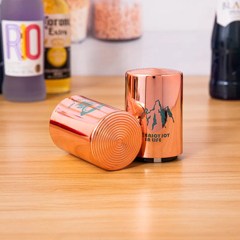 Two copper can openers with robot and mountain designs on a wooden surface, with various bottles in the background.