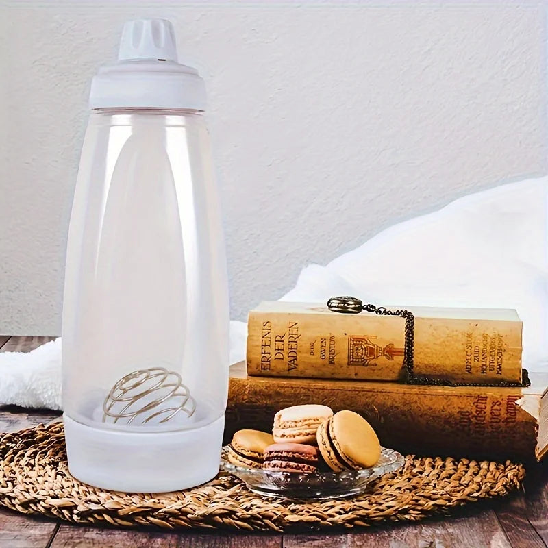 Transparent pancake batter bottle with mixing whisk on table next to books and macarons