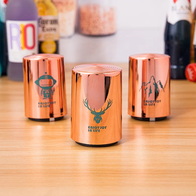Three copper can openers with robot, deer, and mountain designs on a wooden surface with bottles in the background.